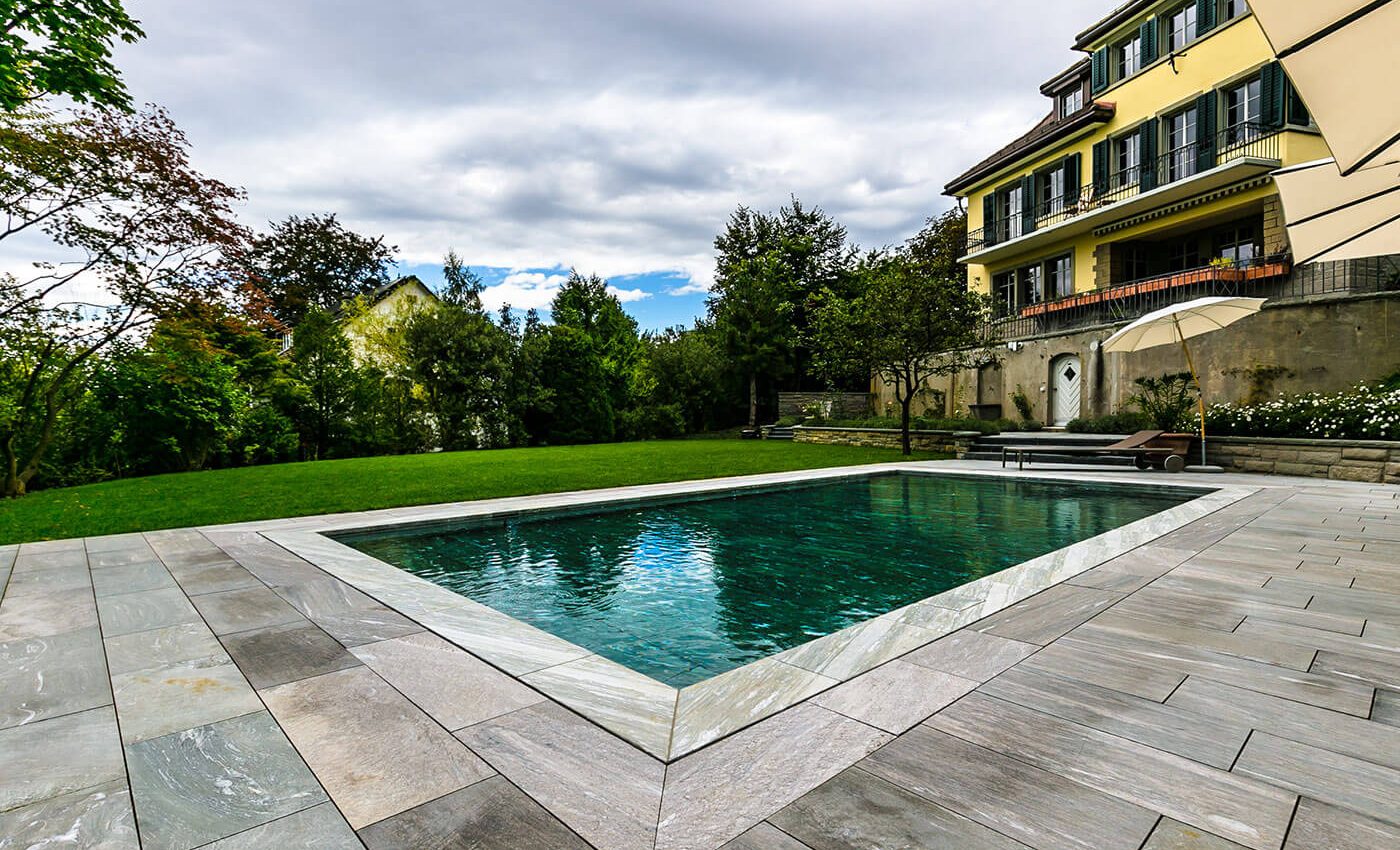 Auf dem Bild ist eine Villa mit einem FlussPool Hubboden zu sehen das mit Granit-Natursteinplatten eingefasst ist.