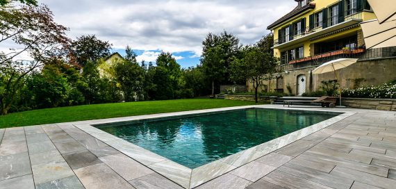 Auf dem Bild ist eine Villa mit einem FlussPool Hubboden zu sehen das mit Granit-Natursteinplatten eingefasst ist.