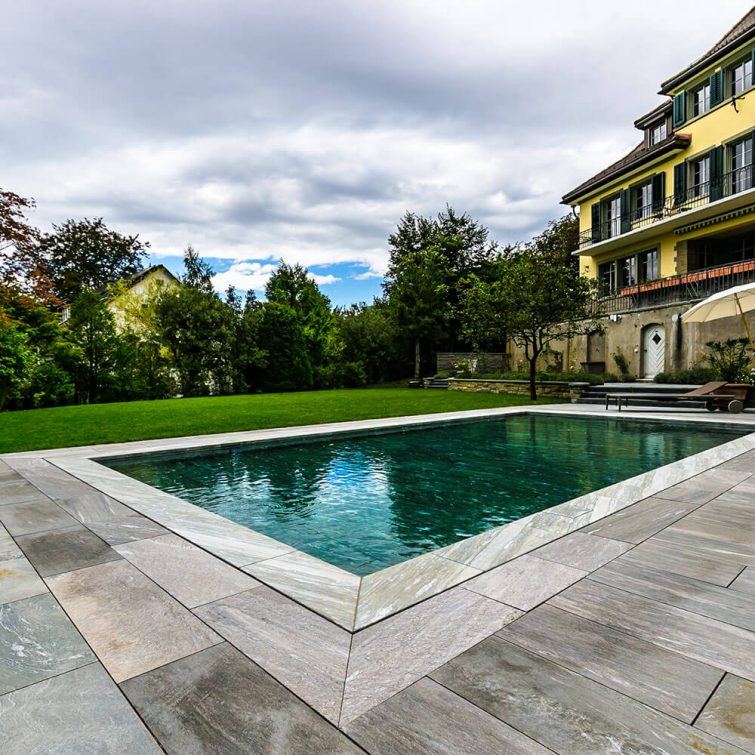 Auf dem Bild ist eine Villa mit einem FlussPool Hubboden zu sehen das mit Granit-Natursteinplatten eingefasst ist.