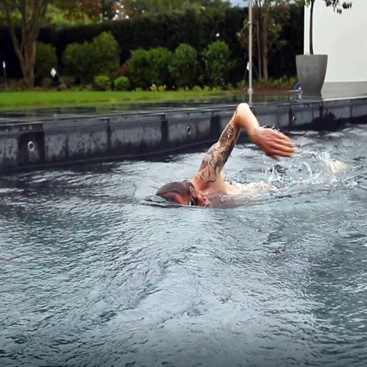 Ein Mann mit Tattoo auf dem Oberarm schwimmt, in seinem Garten, in einem kindersicherem Pool mit Gegenstromanlage von FlussPool in St. Moritz