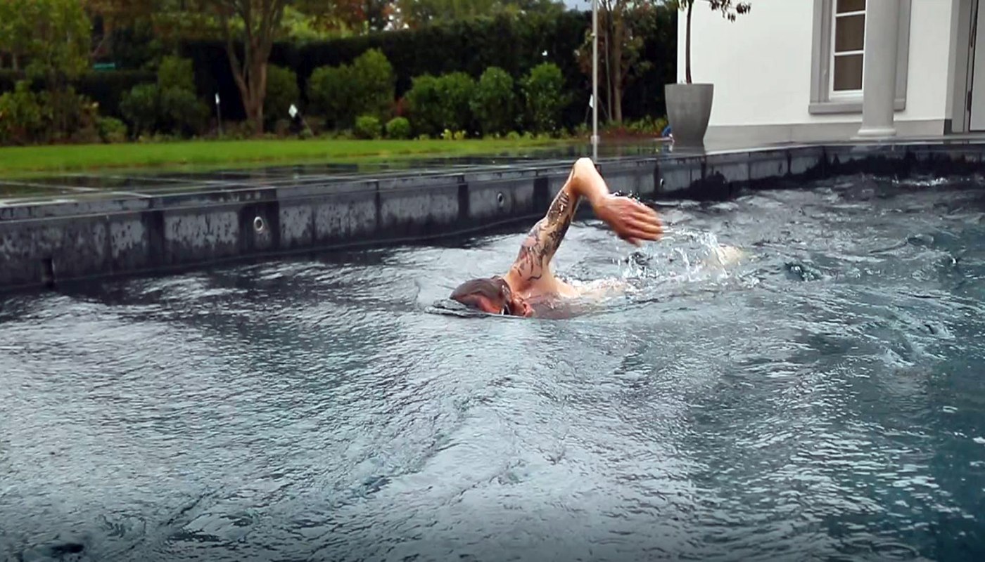 Ein Mann mit Tattoo auf dem Oberarm schwimmt, in seinem Garten, in einem kindersicherem Pool mit Gegenstromanlage von FlussPool in St. Moritz