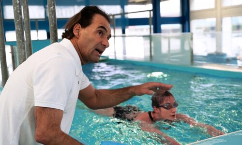 Der Personaltrainer Roy Hinnen, coacht eine Schwimmerin in einem Schwimmkanal. Die Schwimmerin ist im Wasser und Roy Hinnen ist am Beckenrand und erklärt ihr worauf sie achten soll.